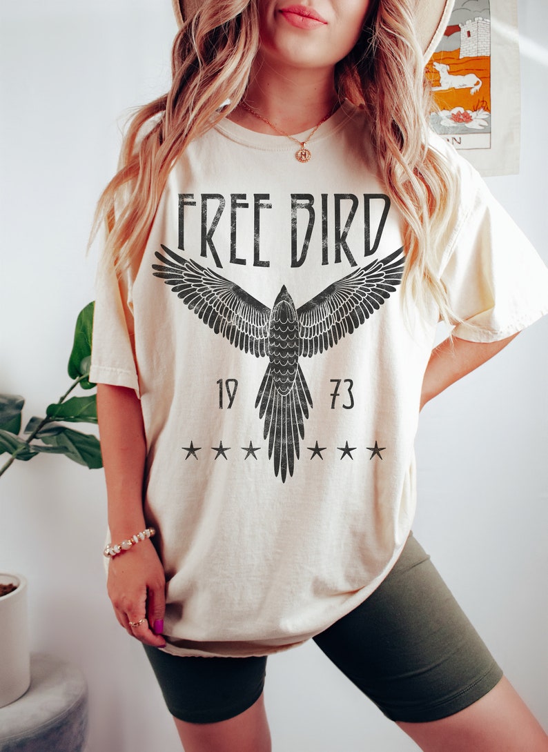 Puede incluir: Una camiseta de color crema con un gr&aacute;fico negro de un p&aacute;jaro con las alas extendidas. El gr&aacute;fico incluye el texto "FREE BIRD" y los n&uacute;meros "19" y "73".
