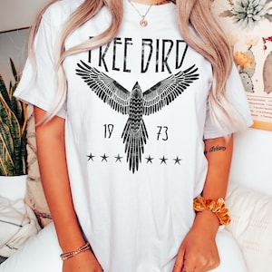 Puede incluir: Camiseta blanca con un gr&aacute;fico negro de un p&aacute;jaro con las alas extendidas y el texto "FREE BIRD" sobre el p&aacute;jaro. Los n&uacute;meros "19" y "73" est&aacute;n a cada lado del p&aacute;jaro, y hay cinco estrellas arriba y abajo del p&aacute;jaro.