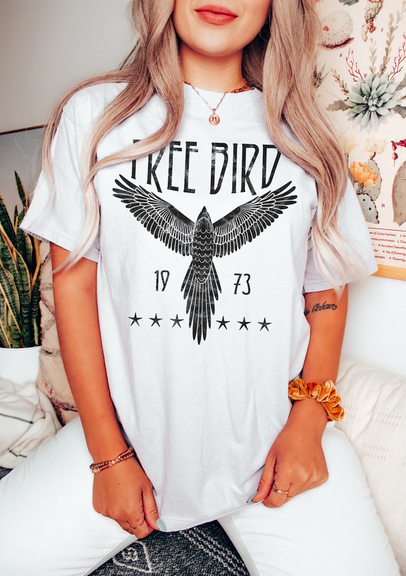 Puede incluir: Camiseta blanca con un gr&aacute;fico negro de un p&aacute;jaro con las alas extendidas y el texto "FREE BIRD" sobre el p&aacute;jaro. Los n&uacute;meros "19" y "73" est&aacute;n a cada lado del p&aacute;jaro, y hay cinco estrellas arriba y abajo del p&aacute;jaro.