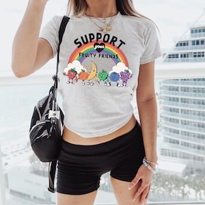 May include: A light grey t-shirt with the slogan "SUPPORT YOUR FRUITY FRIENDS" above a rainbow and cartoon fruits. The fruits include a strawberry, orange, banana, green fruit, blueberry, and grape. The shirt is worn with black shorts.