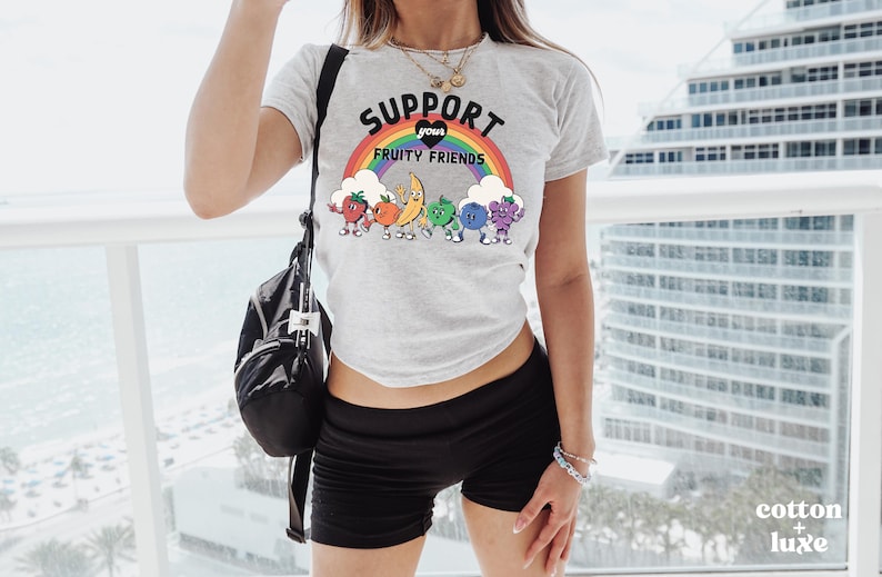 May include: A light grey t-shirt with the slogan "SUPPORT YOUR FRUITY FRIENDS" above a rainbow and cartoon fruits. The fruits include a strawberry, orange, banana, green fruit, blueberry, and grape. The shirt is worn with black shorts.