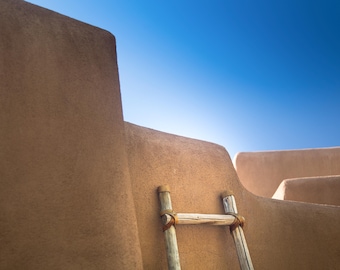 May include: A wooden ladder leans against a tan adobe wall with a blue sky in the background.