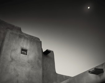 May include: Black and white photograph of a building's exterior against a gradient sky. The building has a textured, stucco-like finish with a small square window. A dark, rectangular element projects from the wall. The moon is visible in the sky.