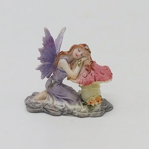 May include: A figurine of a fairy in a lavender dress with purple glitter wings, resting on a pink mushroom. The fairy has red hair and is kneeling on a gray stone base with small flowers.