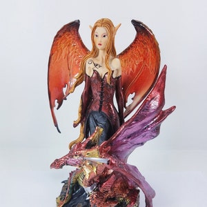 May include: A red and gold dragon figurine with a red and gold fairy figurine standing on top of it. The fairy has long red hair and is wearing a red dress with gold accents. The dragon has large wings and a long tail.
