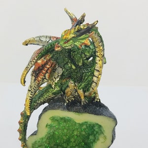 May include: A green, gold, and red dragon figurine perched on a rock with a green crystal inside. The dragon has a long tail and wings.
