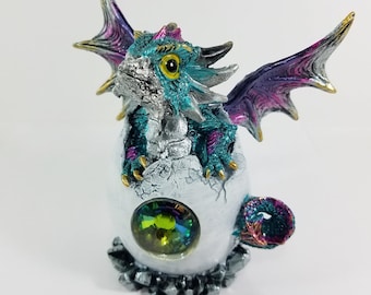 Può includere: Una piccola statuina di drago color turchese e argento con ali viola, appollaiata su un uovo rotto con una gemma verde. Il drago ha un occhio giallo e una coda argentata.