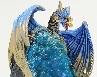 Può includere: Una statuetta di drago blu e oro posata su una roccia con un cristallo blu all'interno. Il drago ha le ali spiegate e la testa girata di lato. La statuetta è realizzata in resina e presenta un design dettagliato.