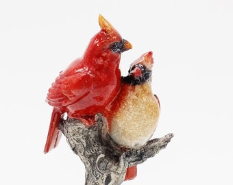 May include: A decorative figurine of two red cardinal birds perched on a tree branch. The male cardinal is a vibrant red, while the female has a tan body. The tree branch and base are textured with naturalistic details.
