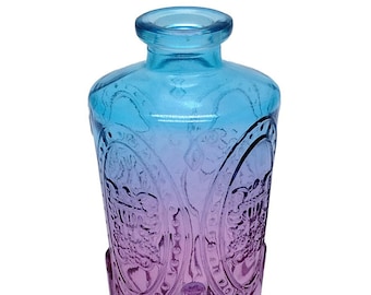 May include: A glass bottle featuring a gradient colour transition from light blue at the neck to purple at the base. The exterior surface is decorated with embossed oval frames containing floral patterns and vertical fluting along the bottom section.