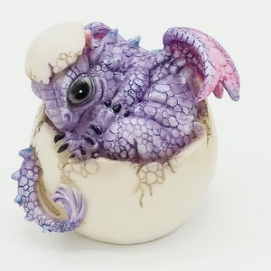 May include: A figurine of a baby dragon hatching from an egg. The dragon is purple with blue and pink accents, and the egg is off-white. The dragon has large, dark eyes and detailed scales. The wings are a soft pink.