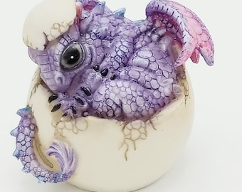 Può includere: Statuetta di un baby drago che esce da un uovo. Il drago è viola con dettagli blu e rosa, l'uovo è bianco sporco. Il drago ha grandi occhi e squame dettagliate. Le ali sono rosa.
