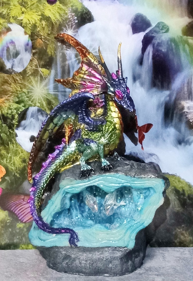 May include: A multi-colored iridescent dragon figurine perched on a rock with a blue crystal cave. The dragon has a long tail and wings, and its eyes are red. The dragon is surrounded by a waterfall and greenery.