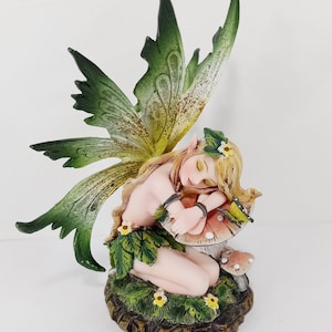 May include: A green and yellow fairy figurine with wings, sleeping on a mushroom with a butterfly on top. The fairy is wearing a green leaf skirt and has a flower crown.
