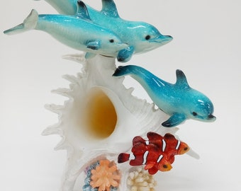 May include: A white seashell with three blue dolphins jumping out of it. There are two orange and red clownfish and coral at the base of the shell.