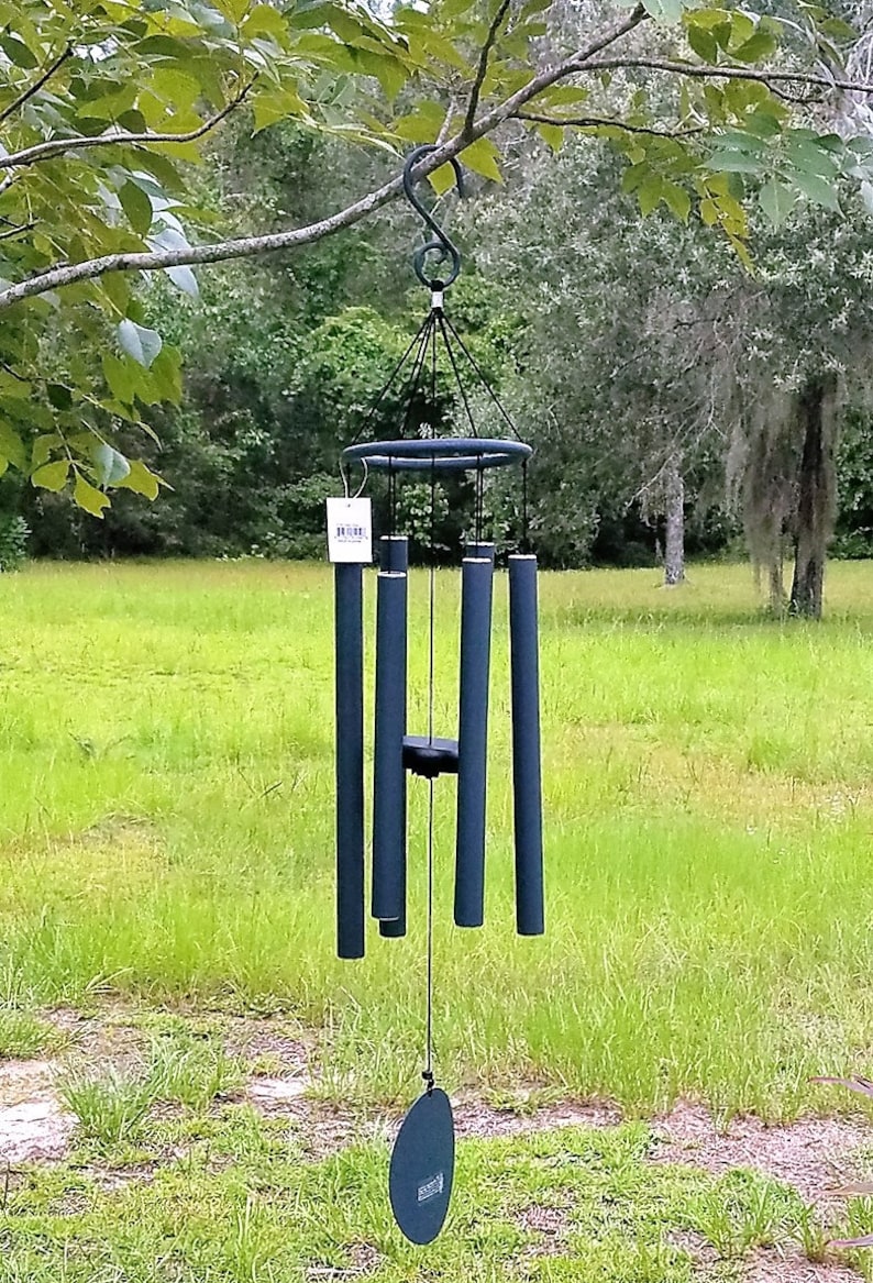 May include: A dark blue wind chime suspended from a tree branch. The chime features a circular top, multiple cylindrical tubes, and a circular wind catcher. The background shows green grass and trees.
