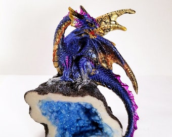 May include: A purple, blue, and gold dragon figurine perched on a rock with a blue crystal inside. The dragon has a long tail and wings.
