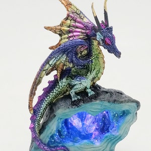 May include: A multi-colored iridescent dragon figurine perched on a rock formation with a glowing blue crystal cave. The dragon has a long tail and wings, and its eyes are red.