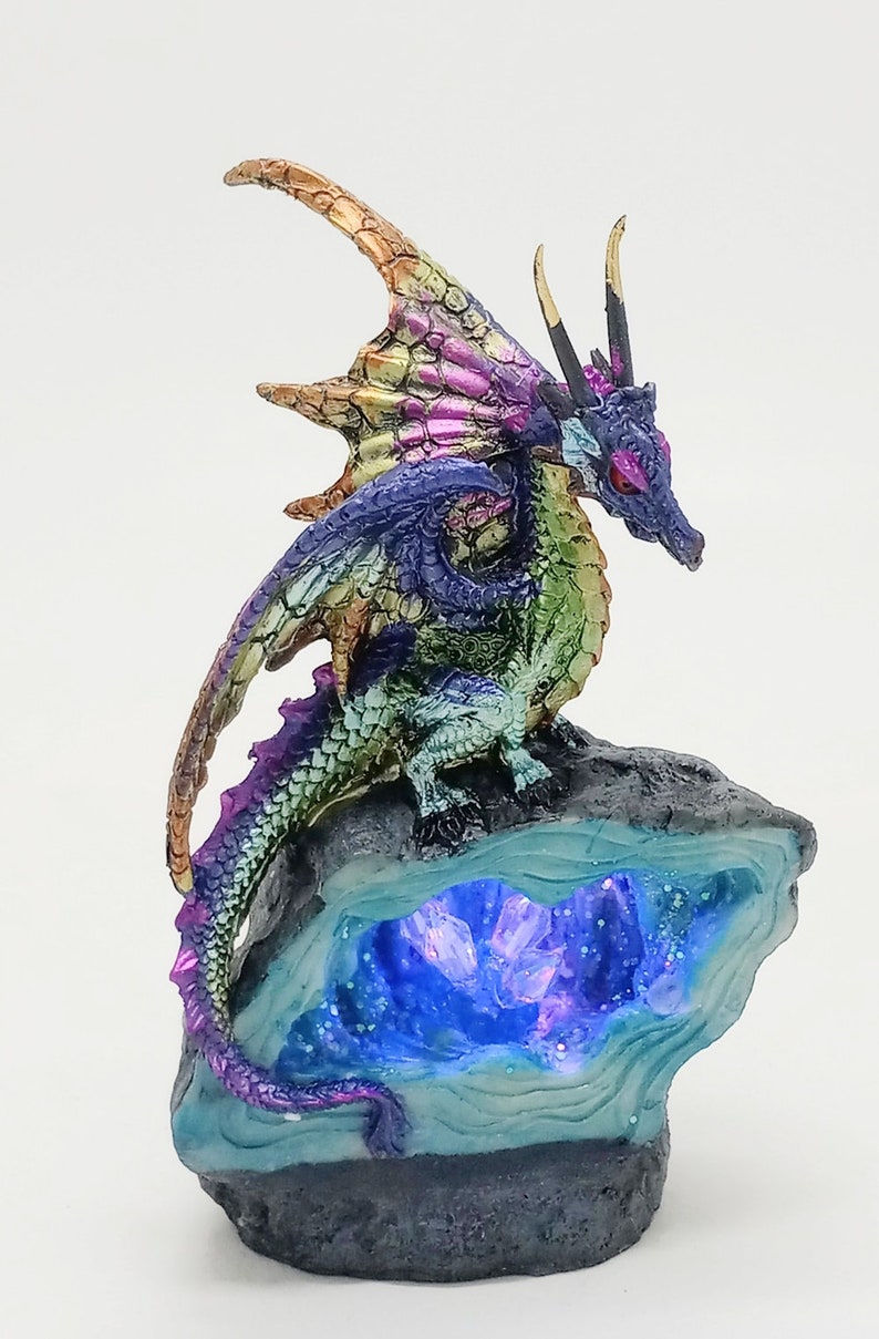 May include: A multi-colored iridescent dragon figurine perched on a rock formation with a glowing blue crystal cave. The dragon has a long tail and wings, and its eyes are red.