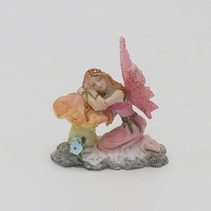 May include: A miniature fairy figurine, featuring a sleeping fairy with long auburn hair, wearing a pink dress, and resting on a mushroom. The fairy has pink wings and is kneeling on a cloud-like base. A small blue flower is visible.