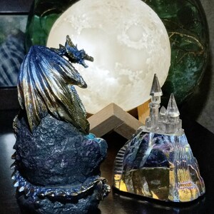K&ouml;nnte beinhalten: Eine blaue und goldene Drachenfigur mit schillernden Fl&uuml;geln, auf einem Felsen sitzend, blickt auf eine Kristallschlossfigur auf einem H&uuml;gel. Der Drache und das Schloss werden von einem leuchtenden Mond beleuchtet.