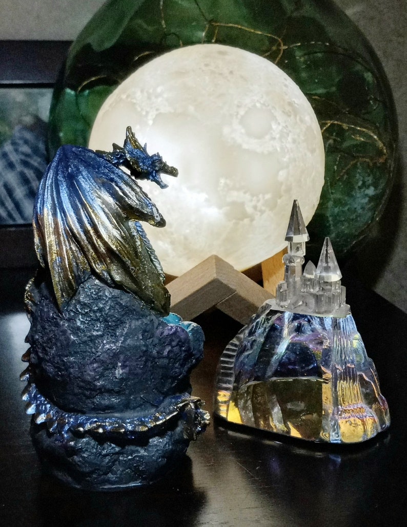 K&ouml;nnte beinhalten: Eine blaue und goldene Drachenfigur mit schillernden Fl&uuml;geln, auf einem Felsen sitzend, blickt auf eine Kristallschlossfigur auf einem H&uuml;gel. Der Drache und das Schloss werden von einem leuchtenden Mond beleuchtet.