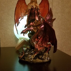May include: A figurine of a female fairy with orange wings riding a red dragon. The fairy is wearing a red and black dress and has long blonde hair. The dragon has gold claws and is standing on a base with rocks and foliage.