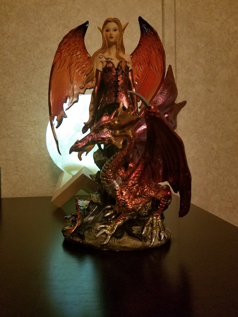 May include: A figurine of a female fairy with orange wings riding a red dragon. The fairy is wearing a red and black dress and has long blonde hair. The dragon has gold claws and is standing on a base with rocks and foliage.