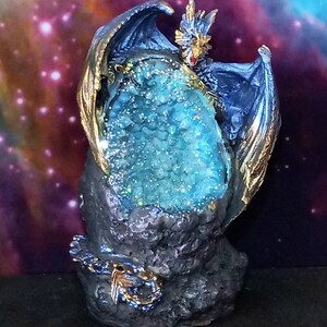 K&ouml;nnte beinhalten: Eine blaue Drachenfigur mit goldenen Akzenten, die eine gro&szlig;e, blaue Kristallgeode h&auml;lt. Der Drache sitzt auf einem schwarzen Felssockel.