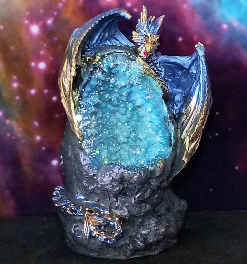 K&ouml;nnte beinhalten: Eine blaue Drachenfigur mit goldenen Akzenten, die eine gro&szlig;e, blaue Kristallgeode h&auml;lt. Der Drache sitzt auf einem schwarzen Felssockel.