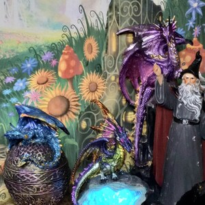 May include: Three dragon figurines, one purple, one green and gold, and one blue and gold. The purple dragon is standing on a rock, the green and gold dragon is standing on a rock with a blue light, and the blue and gold dragon is sitting inside a gold and purple egg.