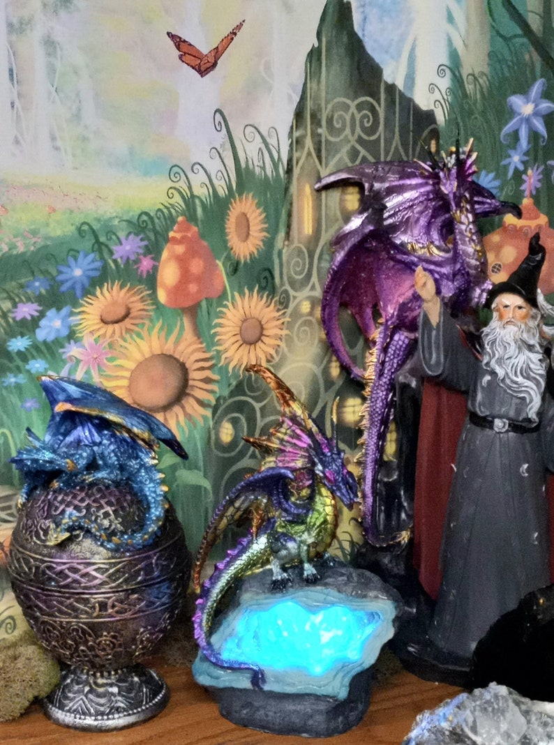 May include: Three dragon figurines, one purple, one green and gold, and one blue and gold. The purple dragon is standing on a rock, the green and gold dragon is standing on a rock with a blue light, and the blue and gold dragon is sitting inside a gold and purple egg.