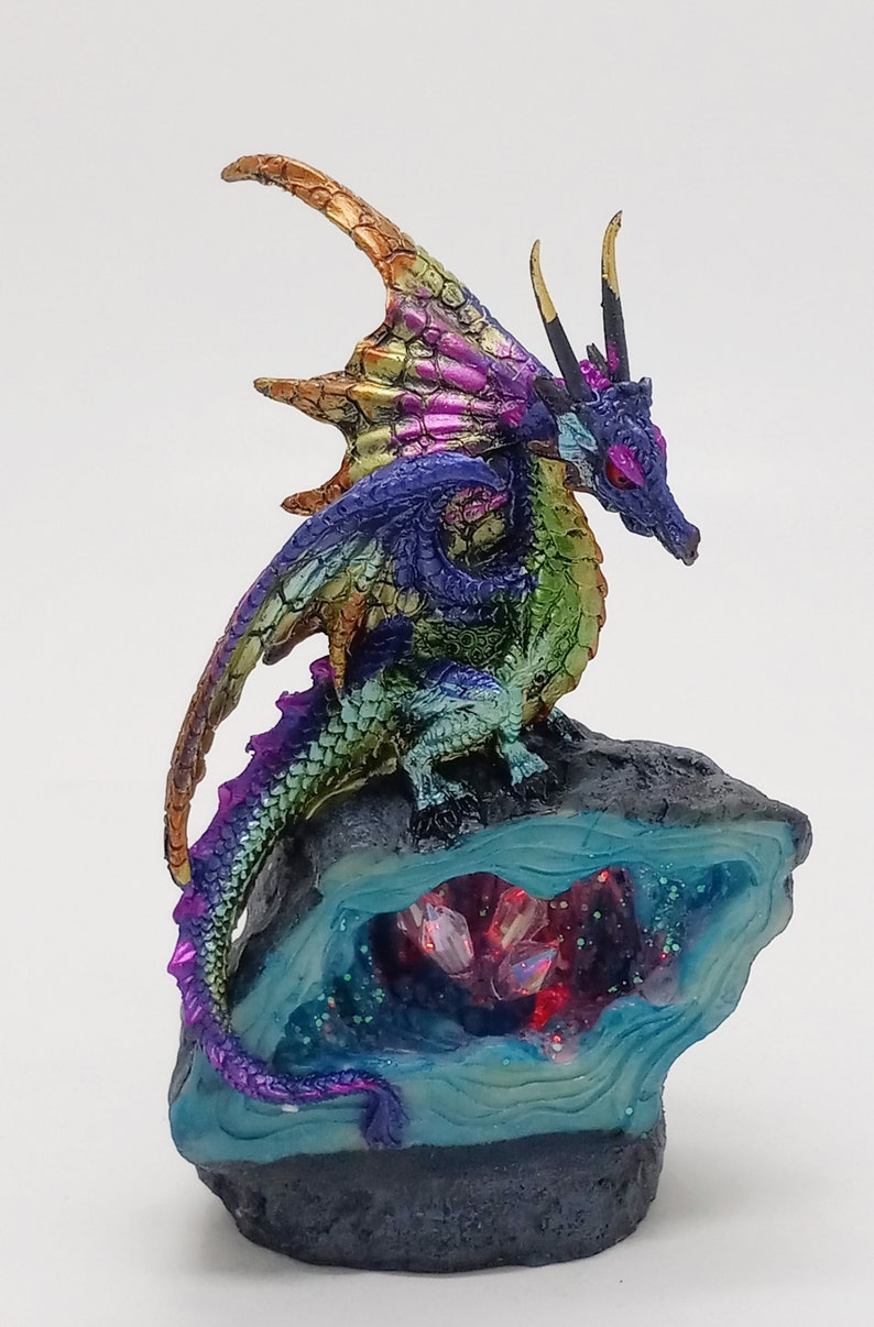 May include: A multi-colored iridescent dragon figurine perched on a rock with a crystal cave. The dragon has a purple tail, green body, and blue wings. The cave is filled with iridescent crystals.