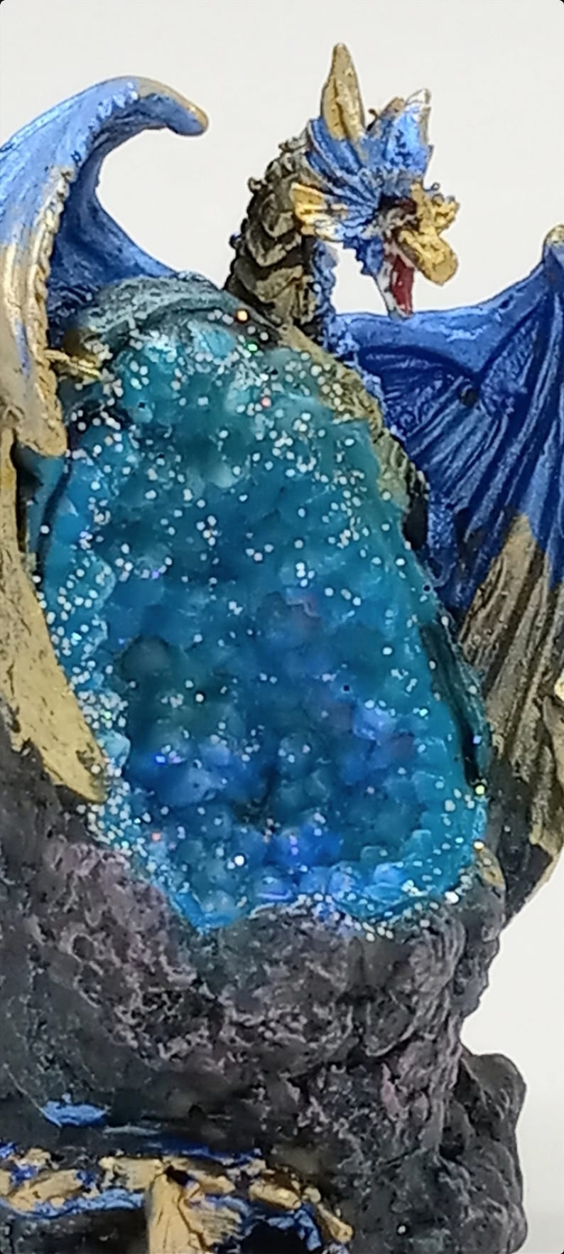 K&ouml;nnte beinhalten: Eine blaue und goldene Drachenfigur mit einem kristallartigen blauen Edelstein, der in seinen R&uuml;cken eingebettet ist. Der Drache sitzt auf einem felsigen Sockel.