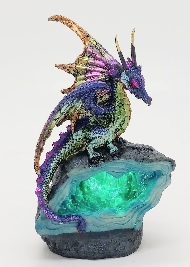 May include: A multi-colored iridescent dragon figurine perched on a rock formation with a glowing green crystal inside. The dragon has a long tail and wings, and its eyes are red.