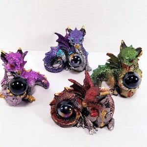 May include: Four small, colorful dragon figurines, each holding a black orb in their paws. The dragons are in various poses, with their wings spread out. The figurines are made of a shiny, metallic material.