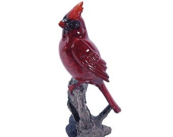 May include: A glossy red cardinal bird figurine with a black face mask and crest, perched on a textured, dark gray tree branch base. The bird has a smooth, reflective finish and is depicted in a side profile view with its tail pointing downward.