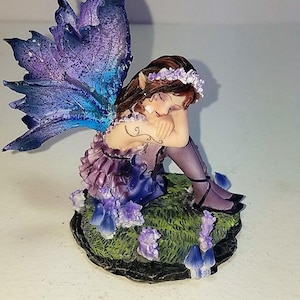 May include: A figurine of a sleeping fairy with brown hair and a floral crown, wearing a purple ruffled dress. The fairy has large, textured blue and purple wings and sits on a green grassy base decorated with small blue mushrooms and flowers.