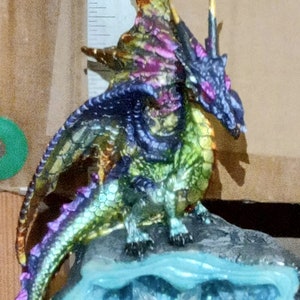 May include: A multi-colored dragon figurine with iridescent scales, perched on a blue and gray rock base with a crystal-like texture. The dragon has a long tail and a horn on its head.