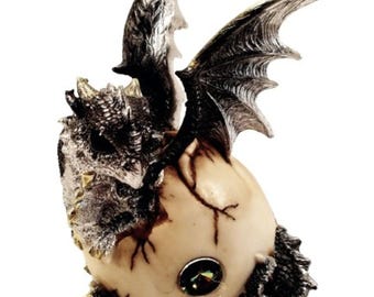 Può includere: Una statuetta di drago argentata e nera che emerge da un uovo incrinato con un occhio di pietra preziosa. Il drago ha le ali spiegate e una superficie dettagliata e testurizzata.