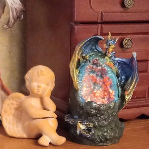 K&ouml;nnte beinhalten: Eine blaue und goldene Drachenfigur mit einem roten und orangefarbenen Kristall im Inneren. Der Drache sitzt auf einem schwarzen Felssockel. Eine kleine, braune Engelsfigur sitzt auf einer Holzoberfl&auml;che.