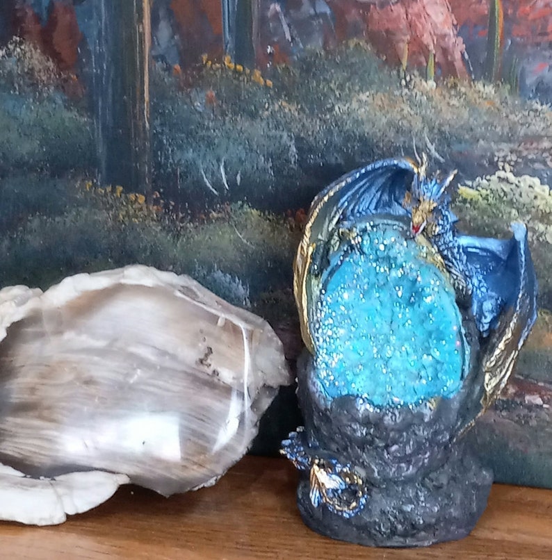 K&ouml;nnte beinhalten: Eine blaue Drachenfigur mit goldenen Akzenten, die sich um einen gro&szlig;en blauen Kristall schlingt. Der Drache sitzt auf einem schwarzen Felssockel.