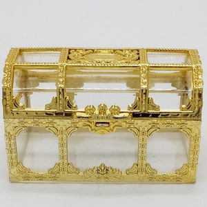 May include: A small, ornate gold-colored treasure chest with a clear glass top. The chest features intricate decorative detailing and a hinged lid. The design includes rectangular and oval shapes, with a clasp closure.