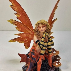 May include: A fairy figurine with orange and yellow wings sits on a rock with a small dragon. The fairy has blonde hair and is wearing a striped outfit.