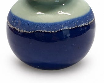 May include: A small, round ceramic vase featuring a glossy finish with a distinct color transition. The upper portion and rim are a pale, muted grey-green, while the lower body is a deep, vibrant blue, separated by a textured, speckled line.