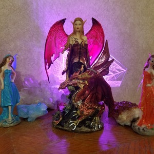 May include: A red and gold dragon figurine with a female fairy figurine riding on its back. The fairy has pink wings and is wearing a red and black dress. The figurine is set against a background of white crystals and a purple light.