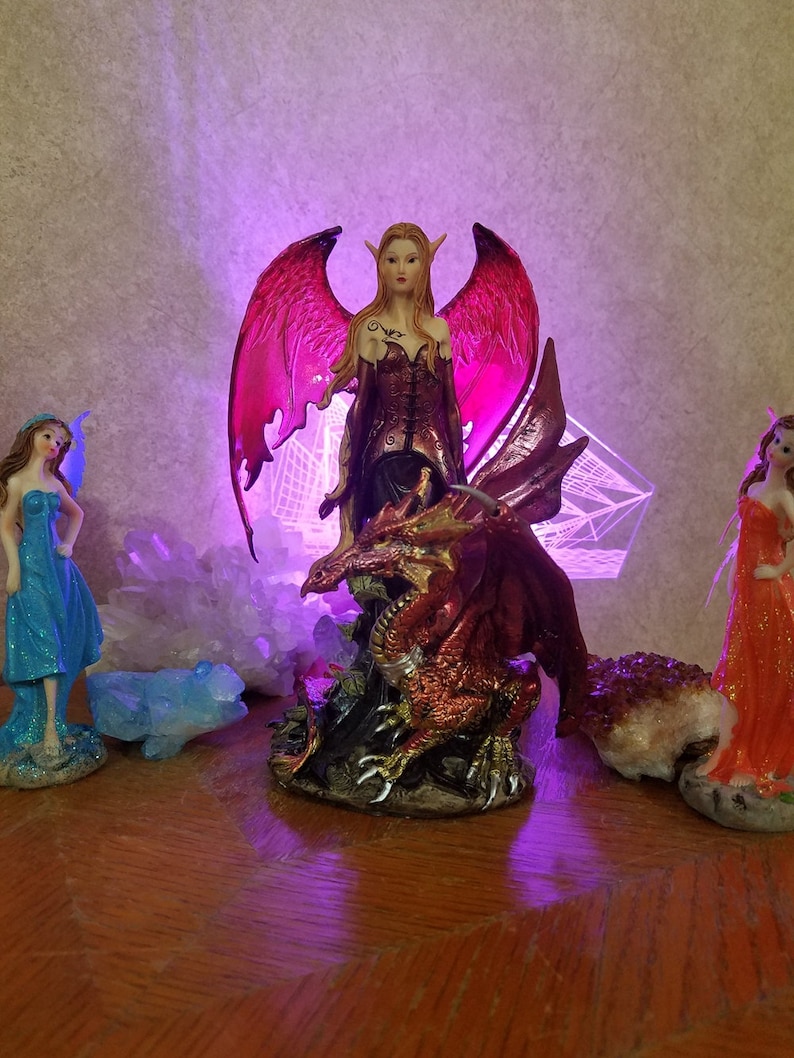 May include: A red and gold dragon figurine with a female fairy figurine riding on its back. The fairy has pink wings and is wearing a red and black dress. The figurine is set against a background of white crystals and a purple light.