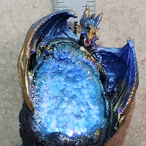 K&ouml;nnte beinhalten: Eine blaue Drachenfigur mit goldenen Akzenten, die eine blaue und wei&szlig;e Kristallkugel h&auml;lt. Der Drache sitzt auf einem schwarzen Felssockel mit einer goldenen Kette.