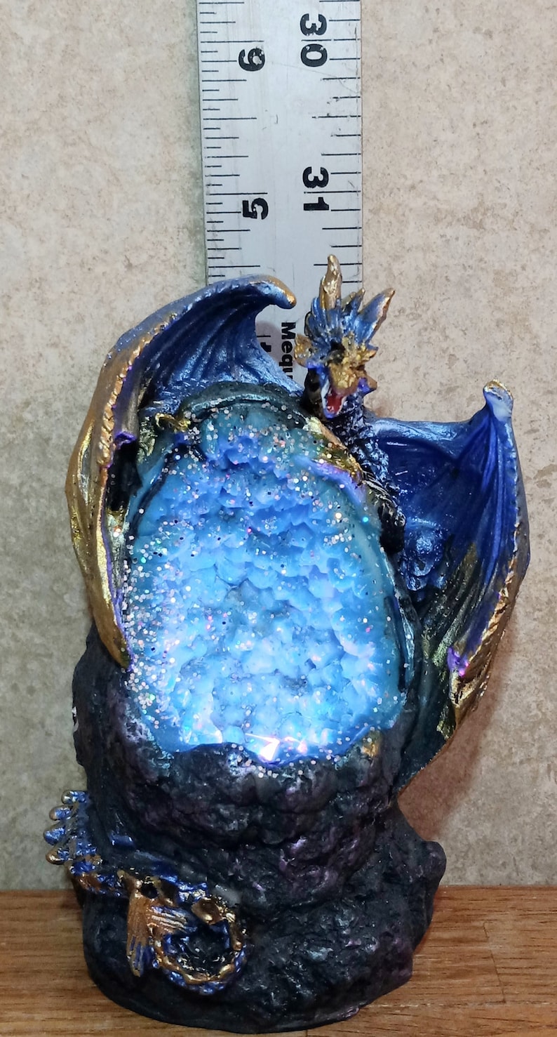 K&ouml;nnte beinhalten: Eine blaue Drachenfigur mit goldenen Akzenten, die eine blaue und wei&szlig;e Kristallkugel h&auml;lt. Der Drache sitzt auf einem schwarzen Felssockel mit einer goldenen Kette.