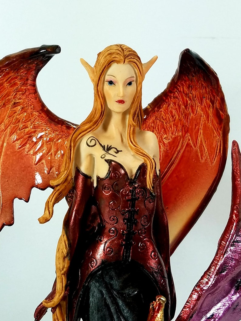 May include: A fantasy figurine of a female with pointed ears, long flowing red hair, and large wings. She is wearing a red and black corset and a long black skirt. The wings are a mix of orange and red.